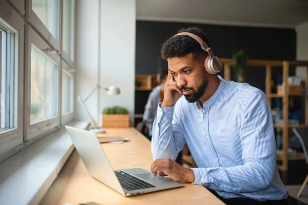 Jovem professor afro-americano com fone de ouvido e laptop dentro de casa na sala de funcionários. — Fotografia de Stock