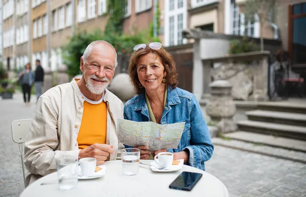 Retrato de feliz casal de idosos turistas sentados e usando mapa ao ar livre na cidade, olhando para a câmera. — Fotografia de Stock