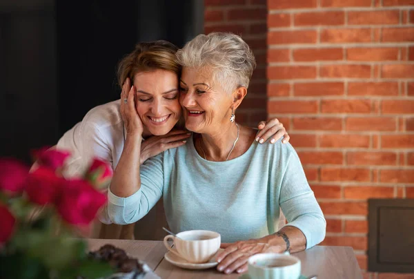 Щаслива старша мати обіймається з дорослою дочкою, сидячи і маючи каву в приміщенні вдома . — стокове фото