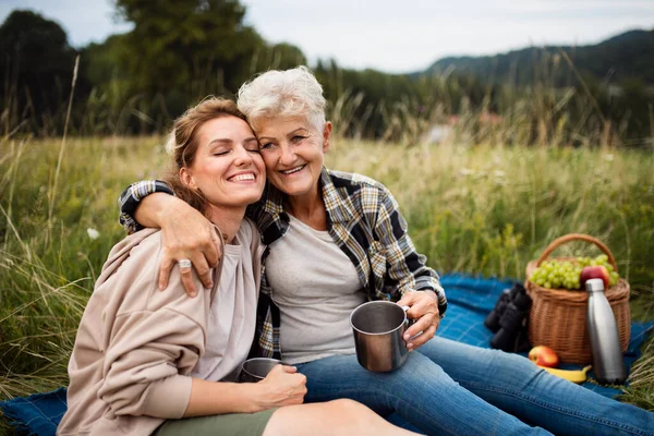 Happy senior mother embracing adult daughter when sitting and having picnic outdoors in nature. — Stock Photo, Image