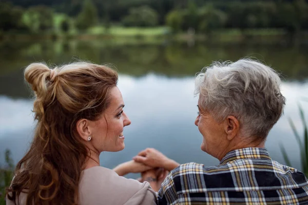 Achteraanzicht van gelukkige senior moeder hand in hand met volwassen dochter wanneer zitten door het meer buiten in de natuur. — Stockfoto