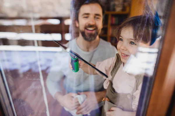 Ragazza felice con padre che pulisce le finestre a casa, concetto di faccende quotidiane. Girato attraverso il vetro. — Foto Stock