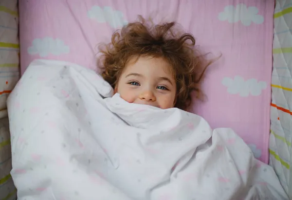 Top view of happy little girl lying in bed hiding under blanket, looking at camera indoors at home — Stock Photo, Image