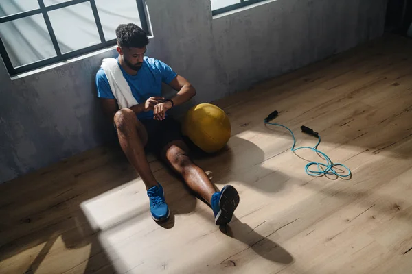Sudut pandang yang tinggi dari pemuda Afrika Amerika olahragawan duduk dan menggunakan smartwatch dalam ruangan di gym. — Stok Foto