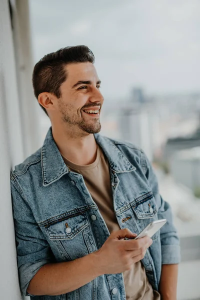 Portret van een gelukkige jonge blanke man met smartphone op het balkon. — Stockfoto