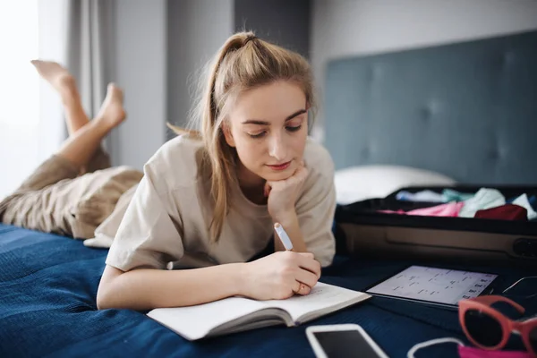 Retrato de mulher jovem escrevendo notas e embalando roupas em mala em casa, planejando férias. — Fotografia de Stock