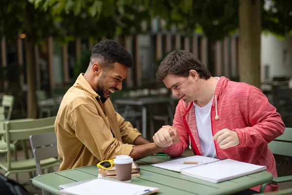 Ung man med Down syndrom med sin mentorskap vän arm brottning utomhus i caféet — Stockfoto