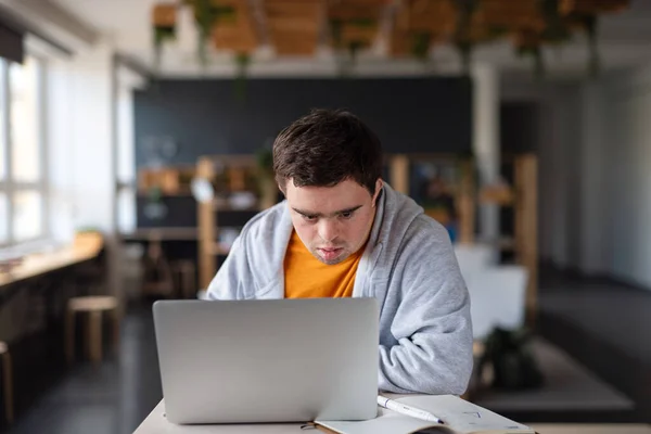 Skoncentrowany młody człowiek z zespołem Downa siedzi i uczy się w szkole, używając laptopa — Zdjęcie stockowe