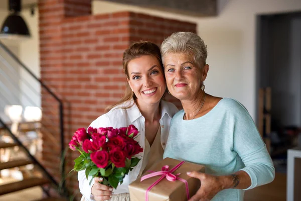 Glückliche Seniorin bekommt Geschenk und Blumenstrauß von erwachsener Tochter zu Hause, schaut in die Kamera. — Stockfoto