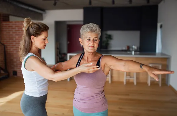 Mujer mayor activa en ropa deportiva haciendo ejercicio con fisioterapeuta femenina en casa. —  Fotos de Stock