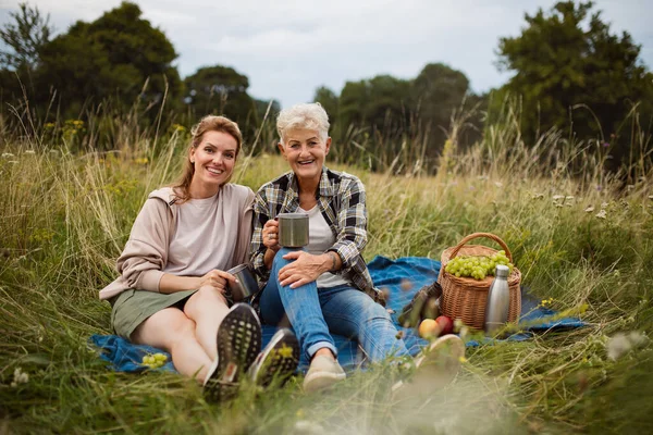 Ältere Mutter und erwachsene Tochter sitzen und picknicken draußen in der Natur und schauen in die Kamera. — Stockfoto