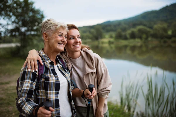 Happy senior mother hiker embracing with adult daughter when looking at lake outdoors in nature — Stock Photo, Image