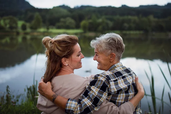 Вид ззаду щасливої старшої матері, що обіймається з дорослою донькою, сидячи на озері на відкритому повітрі в природі — стокове фото