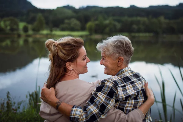 Visão traseira da mãe sênior feliz abraçando com a filha adulta quando sentada no lago ao ar livre na natureza — Fotografia de Stock