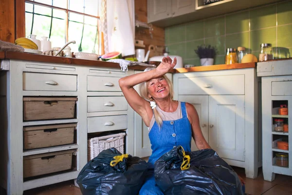 床に座り、キッチンで屋内で廃棄物を分離する疲れシニア女性,持続可能なライフスタイル. — ストック写真