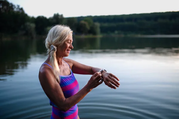 Retrato de la mujer mayor activa nadadora de pie y ajuste smartwatch al aire libre en el lago. — Foto de Stock