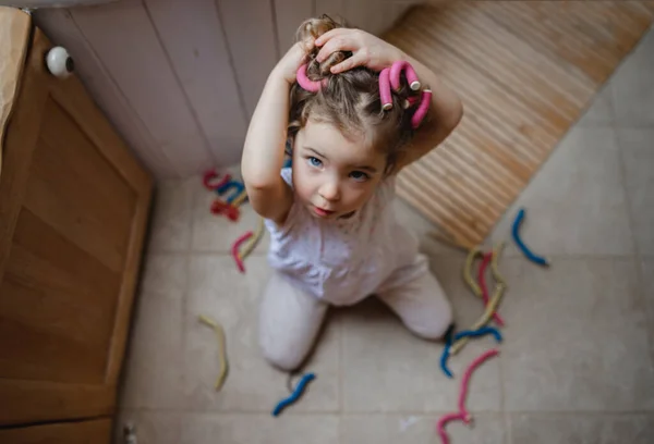 Vista ad alto angolo di bambina che applica bigodini per capelli in casa, guardando la fotocamera. — Foto Stock