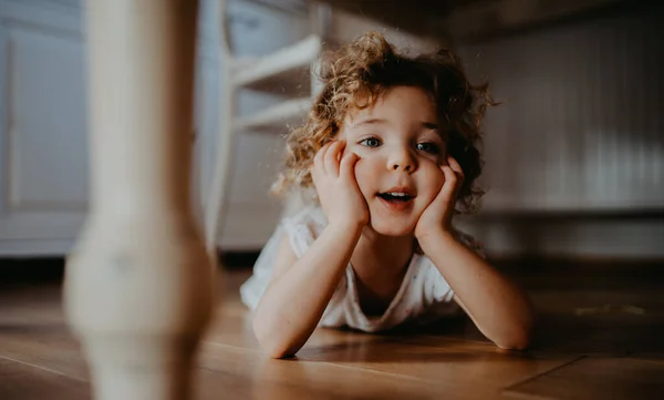 Portrait of cute small girl lying on floor under table indoors at home, looking away. — Stock Photo, Image