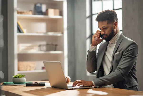 Gelukkig jong Afrikaans Amerikaans zakenman met smartphone werken op een laptop binnen in het kantoor — Stockfoto