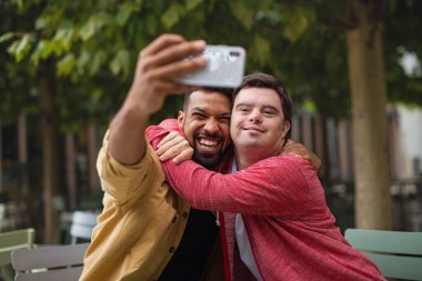 Down sendromlu genç adam ve akıl hocası arkadaşı kafede oturup selfie çekiyorlar.