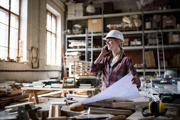 Женщина-инженер держит чертежи и делает телефонный звонок в помещении столярного цеха. — стоковое фото