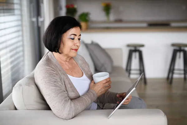 Gelukkig senior vrouw met tablet zitten op de bank binnen thuis, video call concept. — Stockfoto