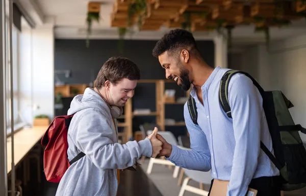 Young happy man with Down syndrome with his mentoring friend greeting indoors at school. — Stock Photo, Image