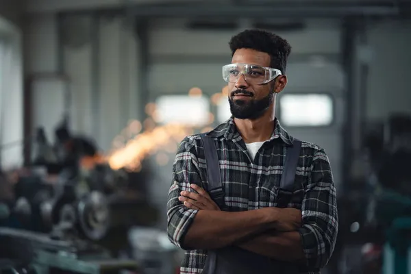 Portrait of young industrial man standing with arms crossed indoors in metal workshop, looking away. — Stock Photo, Image