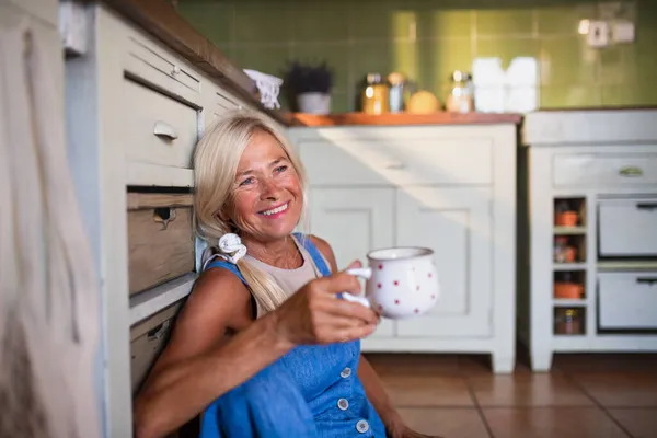 Feliz anciana sentada en el suelo y bebiendo té en la cocina. — Foto de Stock