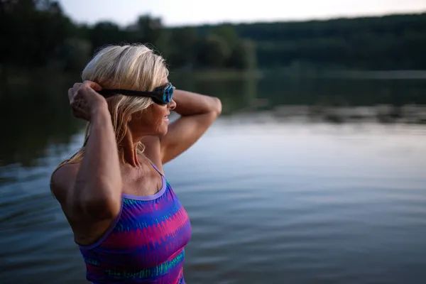 Porträt einer aktiven Seniorenschwimmerin, die im See steht und sich im Freien eine Brille aufsetzt. — Stockfoto