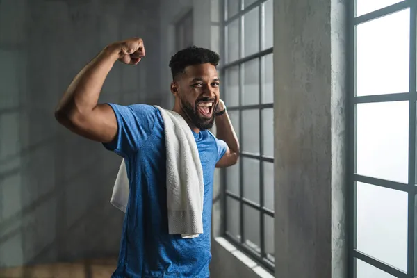 Счастливый молодой афроамериканский спортсмен, стоящий в помещении в спортзале, смотрящий в камеру, демонстрирующий мускулы — стоковое фото