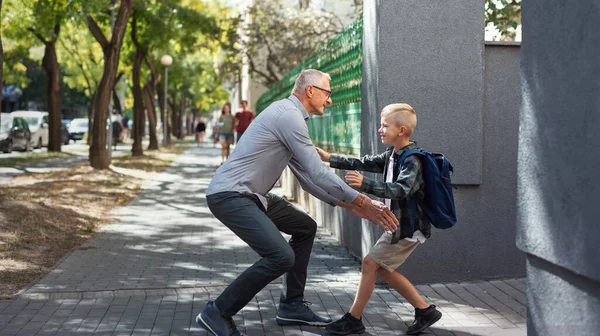 Feliz estudiante corriendo hacia su abuelo esperándolo después de la escuela al aire libre en la calle. — Foto de Stock