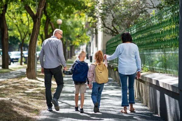 Vista trasera de los abuelos que llevan a sus nietos a casa desde la escuela, caminando al aire libre en la calle. — Foto de Stock