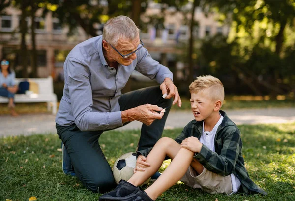 Malý chlapec se zraněnou nohou pláče, jeho dědeček mu dává sádru venku v parku. — Stock fotografie