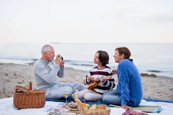 Happy senior couple with granddaughter sitting on blanket and having picnic outdoors on beach by sea. — Stock Photo, Image