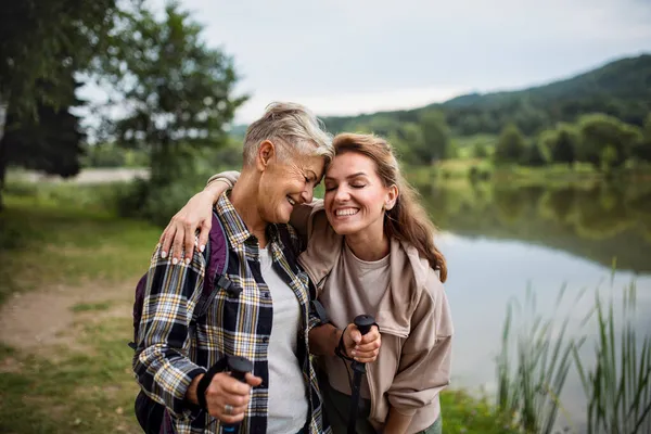 Happy senior mother hiker embracing with adult daughter by lake outdoors in nature — Stock Photo, Image