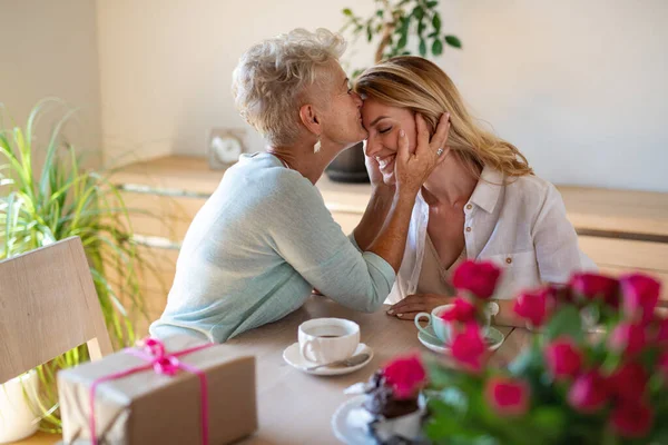 Bonne mère aînée prenant un café avec une fille adulte à l'intérieur à la maison, l'embrassant sur le front. — Photo