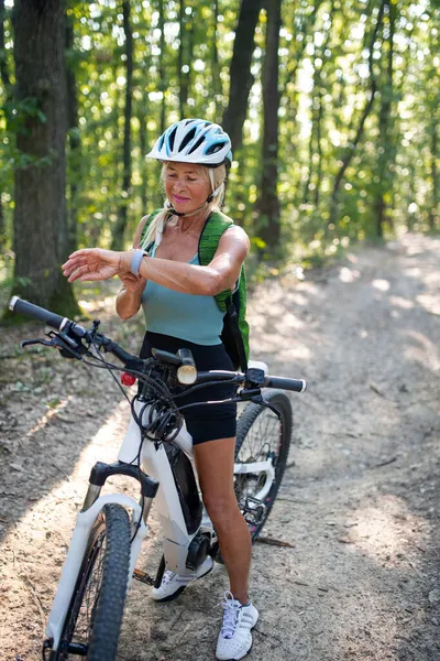 Aktywna starsza kobieta rowerzystka siedzi na rowerze i ustawia smartwatch na zewnątrz w lesie. — Zdjęcie stockowe