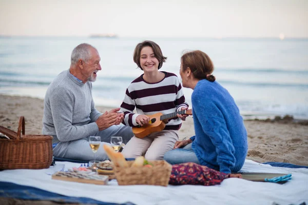 Šťastný starší pár s vnučkou sedí na dece a mají piknik venku na pláži u moře. — Stock fotografie