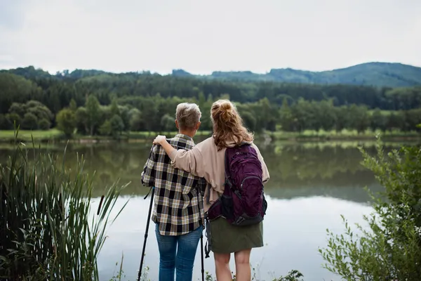 Rear view of senior mother embracing with adult daughter when standing by lake outdoors in nature — Stock Photo, Image