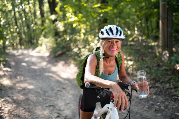 Happy active senior kobieta rowerzysta trzymając butelkę wody podczas siedzenia na rowerze na świeżym powietrzu w lesie. — Zdjęcie stockowe