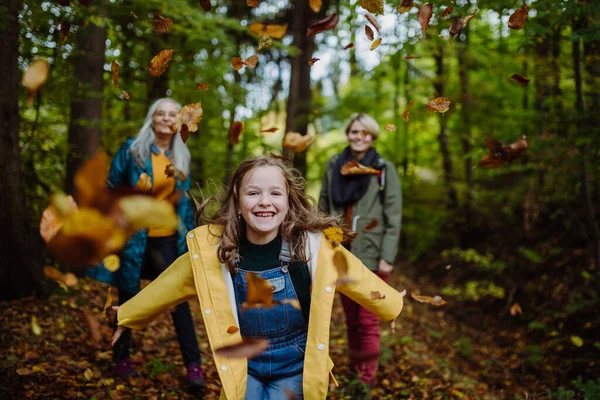 Mutlu küçük kız sonbahar yürüyüşü sırasında annesi ve büyükannesiyle ormanda koşup yaprak fırlatıyor. — Stok fotoğraf