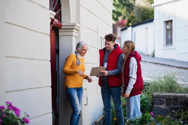 Young door to door volunteers talking to senior woman and taking survey at her front door. — Stock Photo, Image