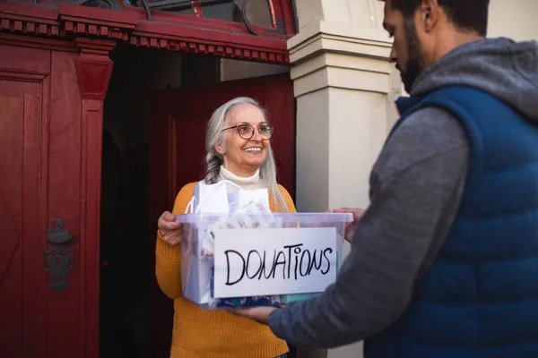 Jeune bénévole donnant boîte de don à une femme âgée debout dans l'embrasure de porte, concept social et caritatif — Photo