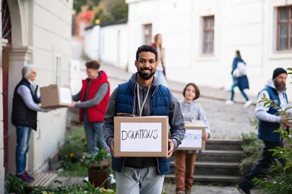 Happy young volunteer with team delivering donation boxes and looking at camera, social care concept — Stock Photo, Image