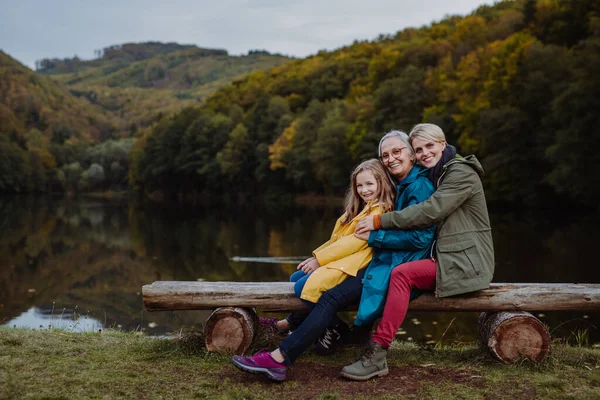 Маленькая девочка с мамой и бабушкой, сидящая на скамейке и смотрящая на фотоаппарат у озера. — стоковое фото