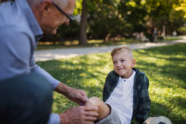 Маленький мальчик с травмой ноги получает штукатурку от дедушки на открытом воздухе в парке. — стоковое фото