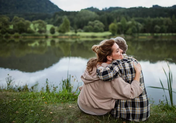 Rückansicht einer glücklichen Seniorin, die sich mit ihrer erwachsenen Tochter umarmt, wenn sie in der Natur am See sitzt — Stockfoto