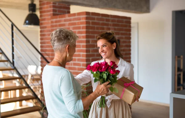 Šťastná dospělá dcera přinášející dárek a kytice pro starší matku doma. — Stock fotografie