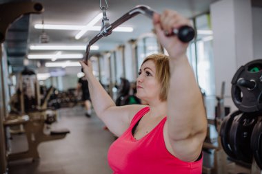 Orta yaşlı ve beden ölçülerinde bir kadın spor salonunda, kapalı bir yerde, bir çekme makinesinde çalışıyor.